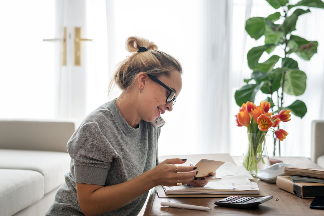 Blonde girl with black glasses in grey sweatshirt sitting at desk on the phone in an airy living room with orange tulips and green house plant