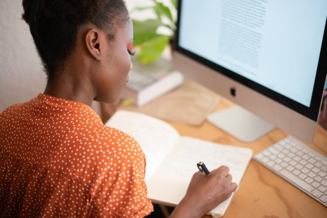 Young Black woman in orange polka dot shirt sitting at a desk in front of a desktop monitor and writing in a notebook